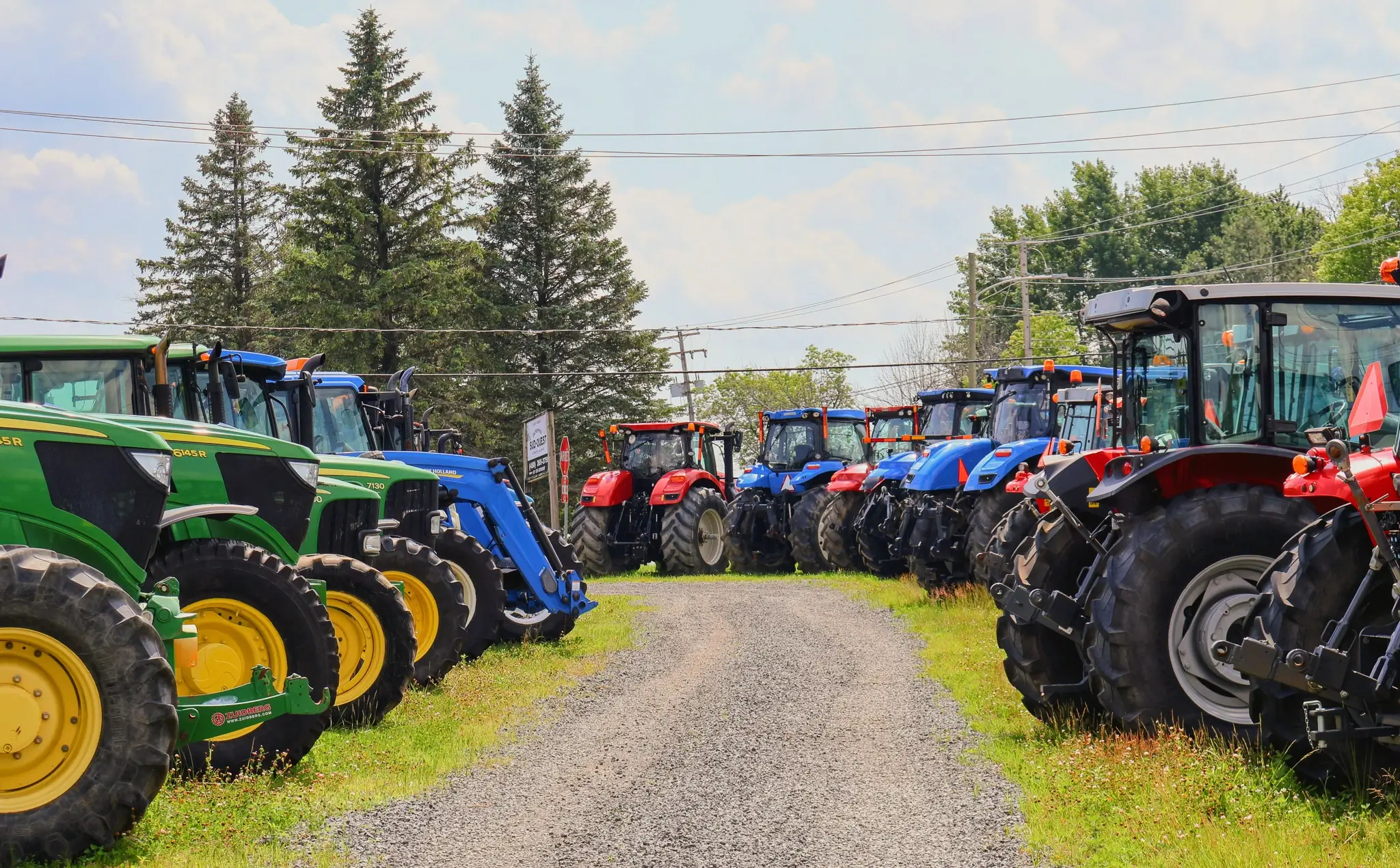Large selection of farm tractors displayed on a dealership lot along a gravel access road.