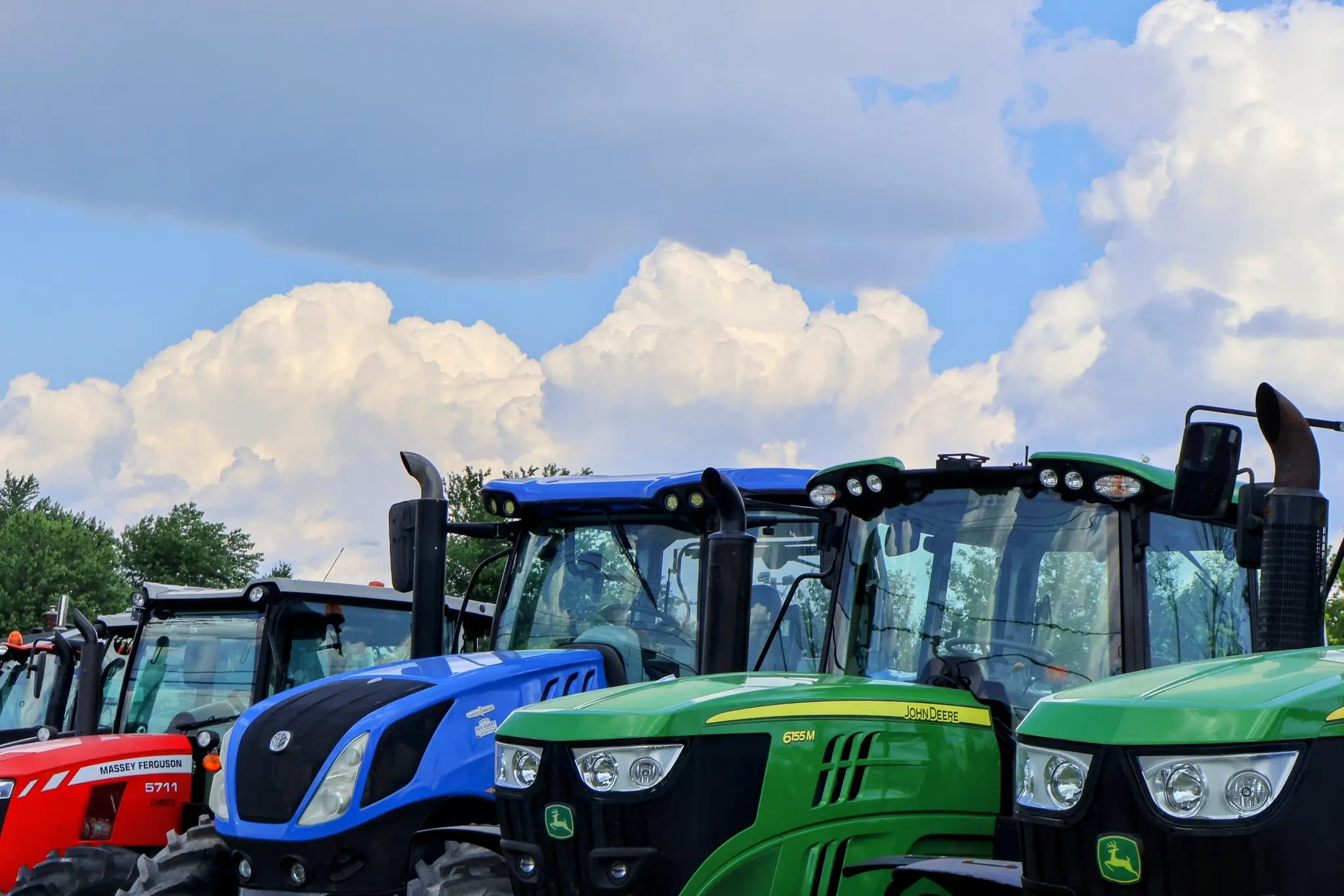 Front view of multiple tractors in different colors lined up outdoors beneath large white clouds.